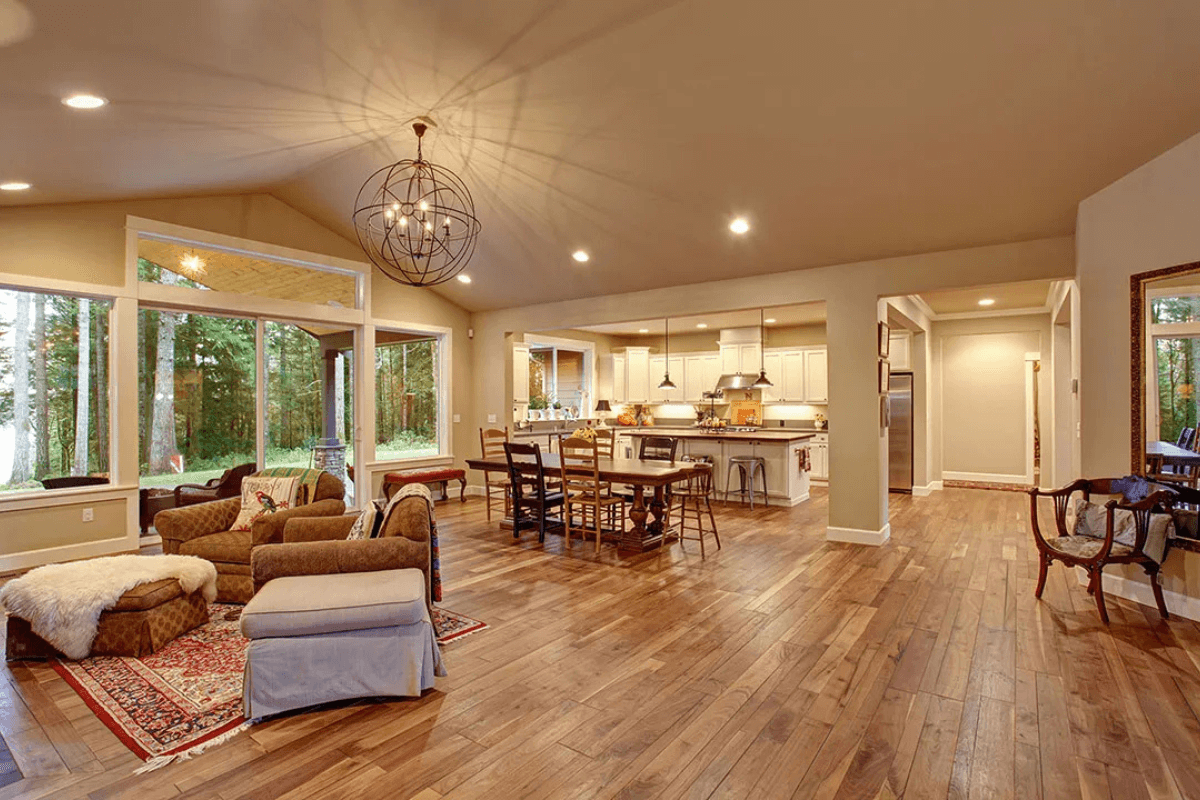 A cozy living room with big windows showing trees outside, wooden floors, comfy chairs, and a dining table in the back