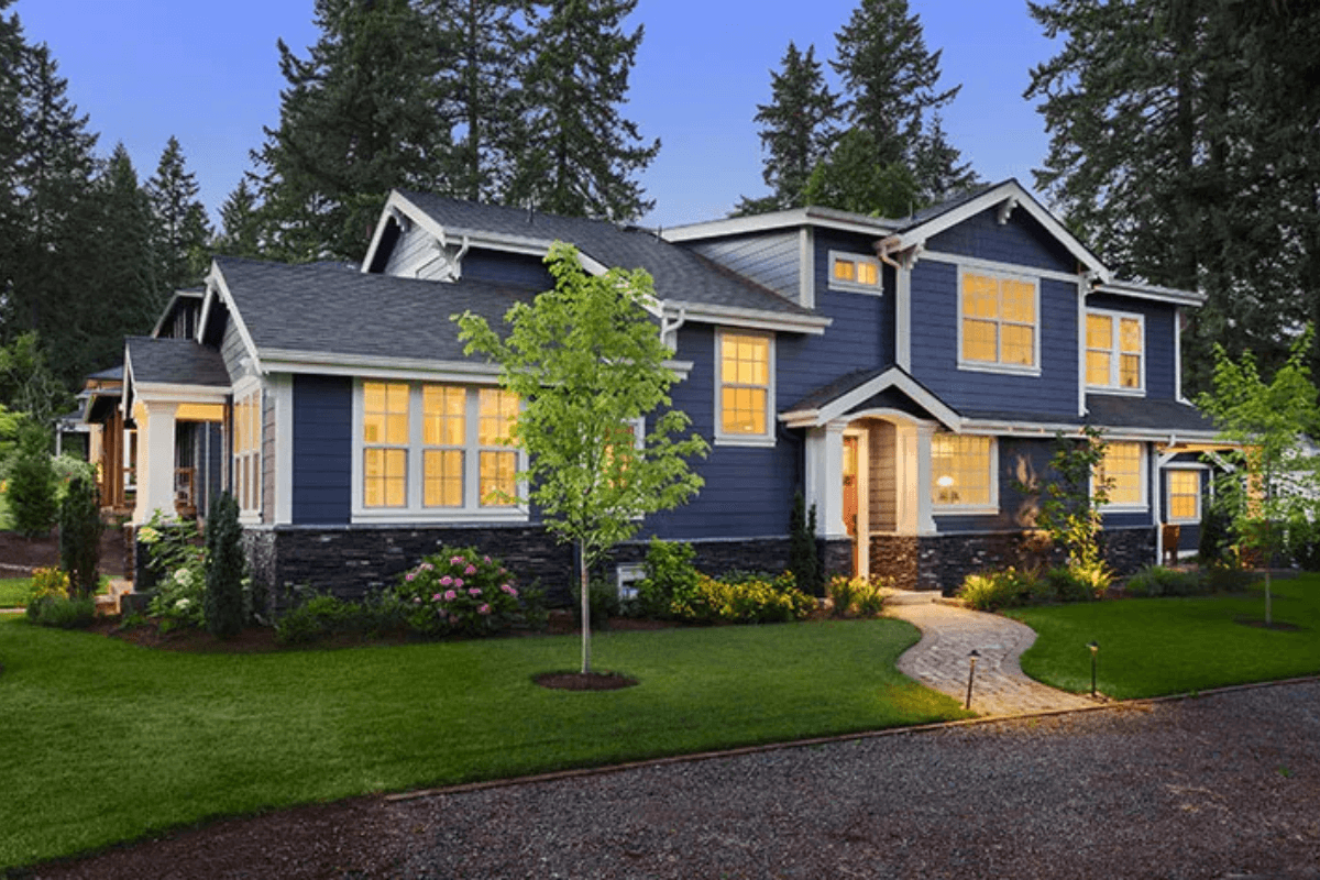 A big blue house with bright yellow lights inside, green grass, and tall trees around it.