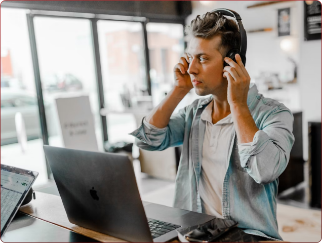 Young man wearing headphones and adjusting them while working on a laptop at a modern cafe with large windows in the background.