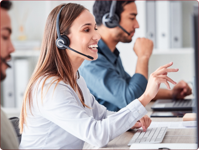 Smiling female customer service representative wearing a headset, pointing at a computer screen while working in an office alongside colleagues.