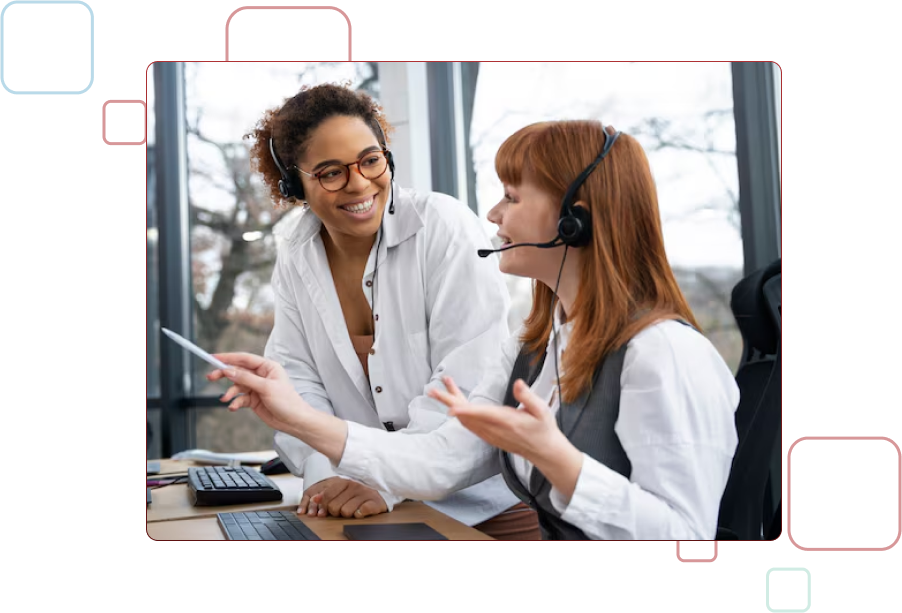 Smiling female customer service representative wearing a headset, pointing at a computer screen while working in an office alongside colleagues.