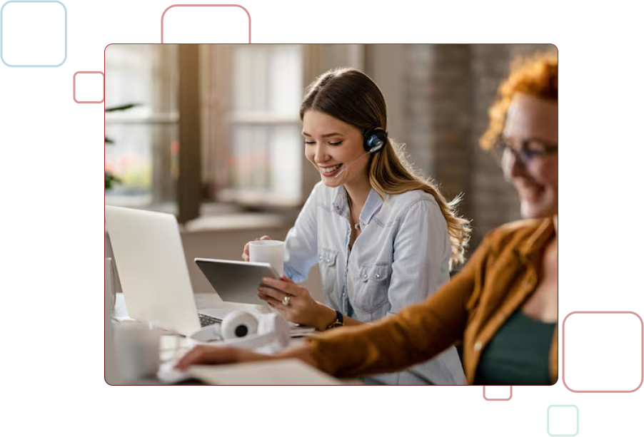 Smiling female customer service representative wearing a headset, pointing at a computer screen while working in an office alongside colleagues.